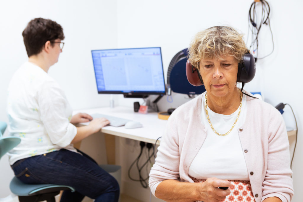A lady doing a hearing test with the audiologist behind her conducting the hearing test using the computer
