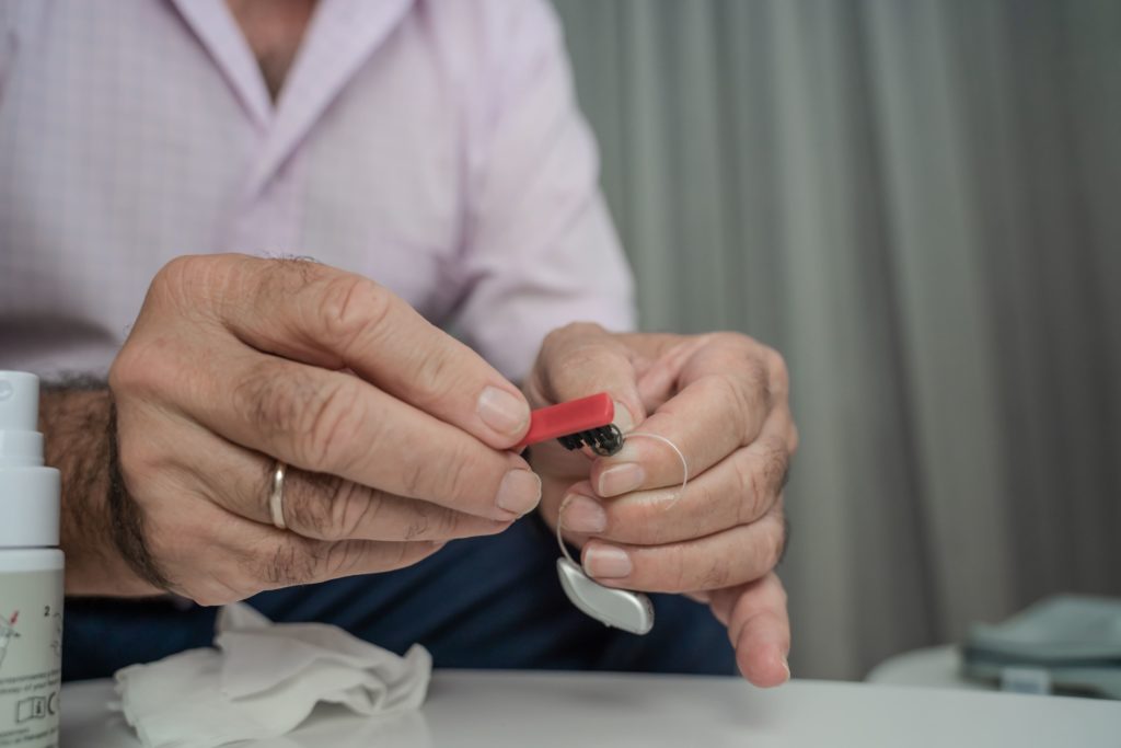 A man is sitting at a table and cleaning his receiver-in-canal hearing aid with a small brush and a cloth.