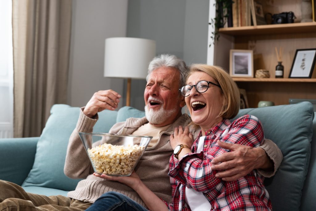 A couple are sitting on the sofa eating popcorn while watching a film and laughing.