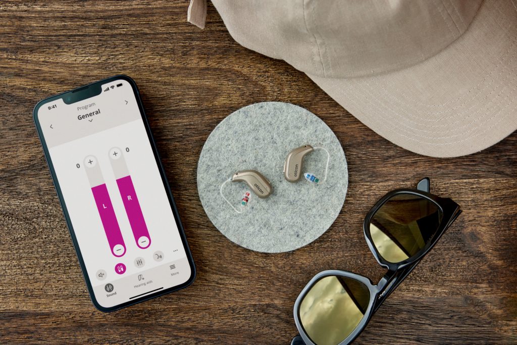 A pair of Oticon Intent hearing aids are laying on a table with a pair of sunglasses, an iPhone and a cap.