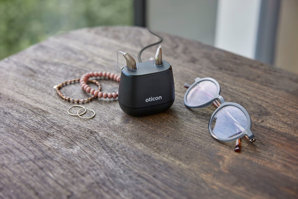 A pair of Oticon Intent hearing aids are in their charger on a table, with a pair of sunglasses, a pair of earings and 2 bracelets next to them.